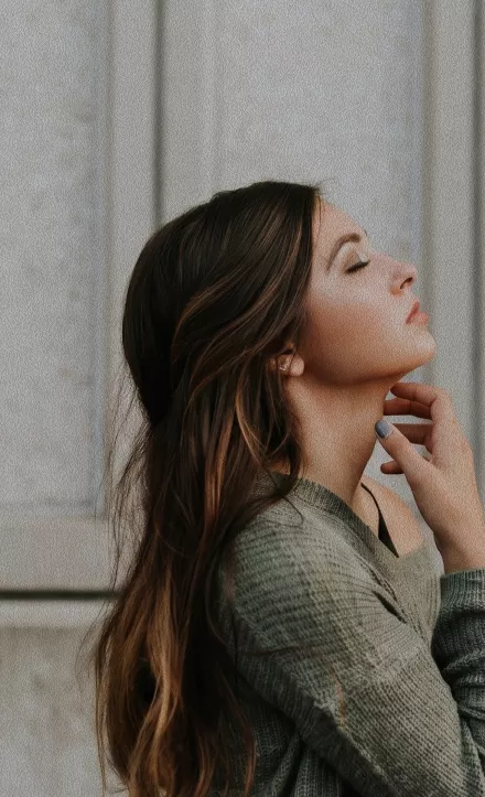 A side profile of a woman with long brown hair and closed eyes tilting her head back against a stone wall.