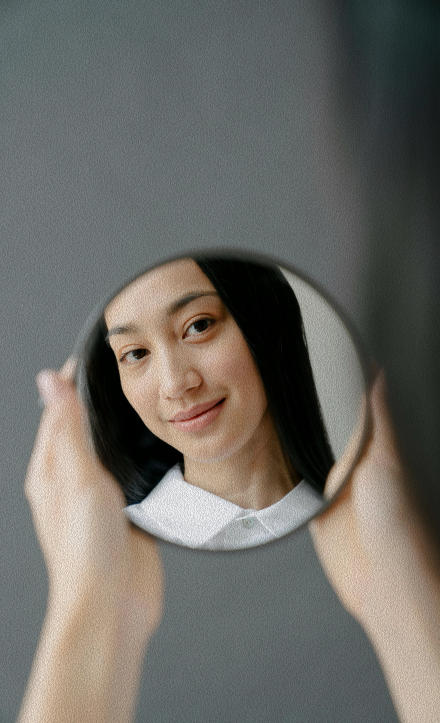 A woman with black hair smiling softly at her reflection in a small round handheld mirror against a gray wall.