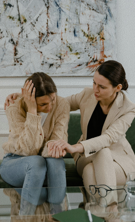 A therapist in a beige blazer offering comfort to a distressed woman holding her head in an office setting.