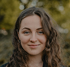 A young woman with curly brown hair smiling softly outdoors with warm blurred autumn foliage in the background.