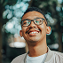 Close-up portrait of a smiling young man with glasses and a mustache, positioned against a blurred greenery background.