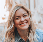 A cheerful woman with blonde wavy hair wearing a denim jacket smiling outdoors with a blurred stone background.